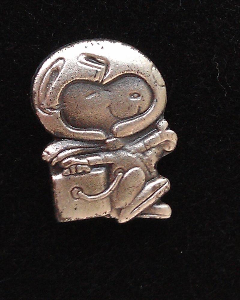 A small silver Snoopy lapel pin in a spacesuit and helmet, photographed on a plain background